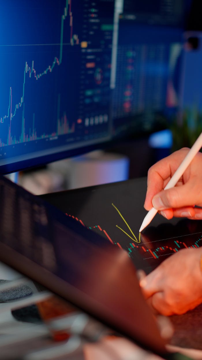 Services Person using a stylus on a tablet for stock market analysis, with charts on a monitor.